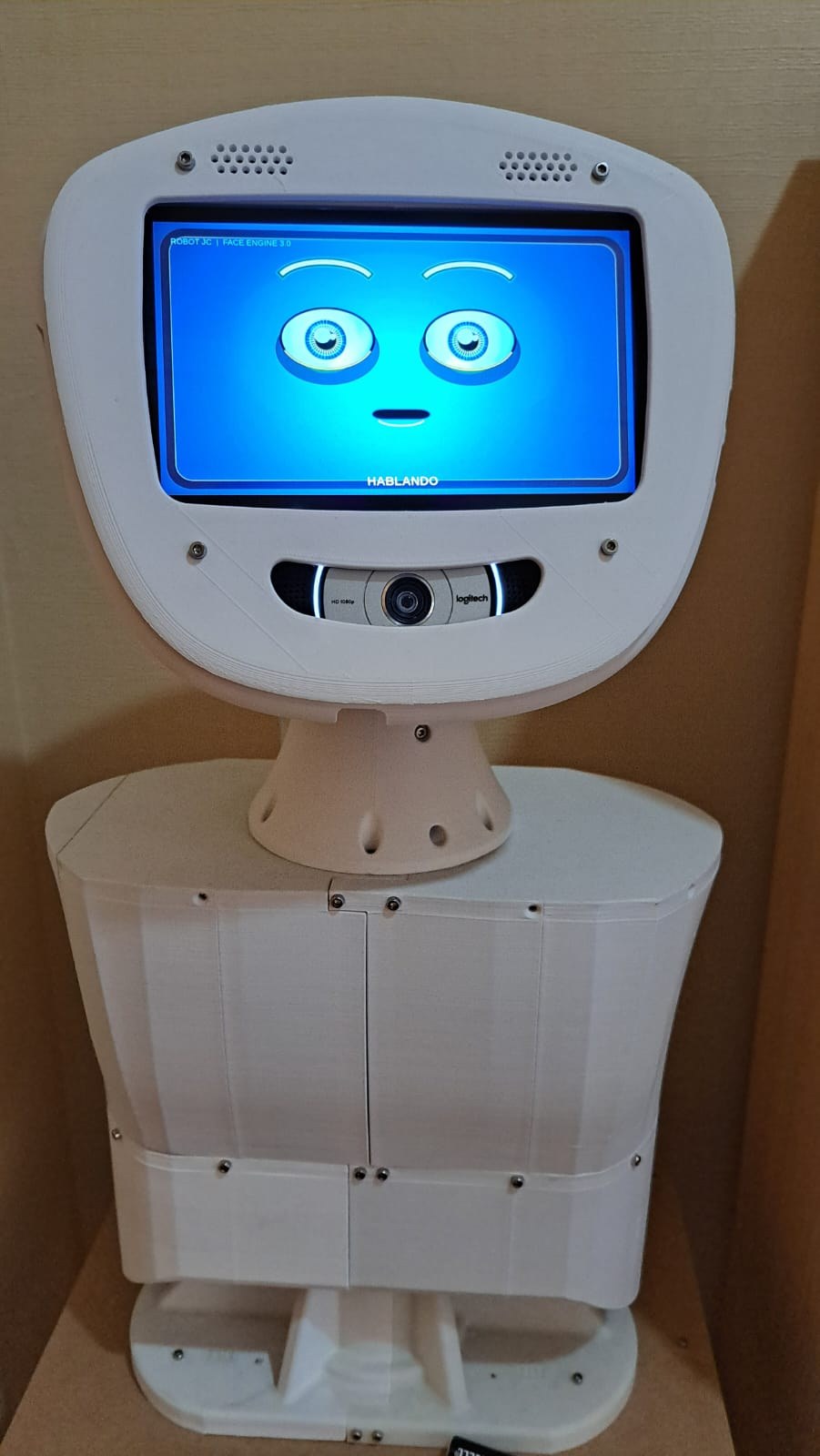 Robot JC de frente, con su rostro digital azul, cámara y estructura física blanca